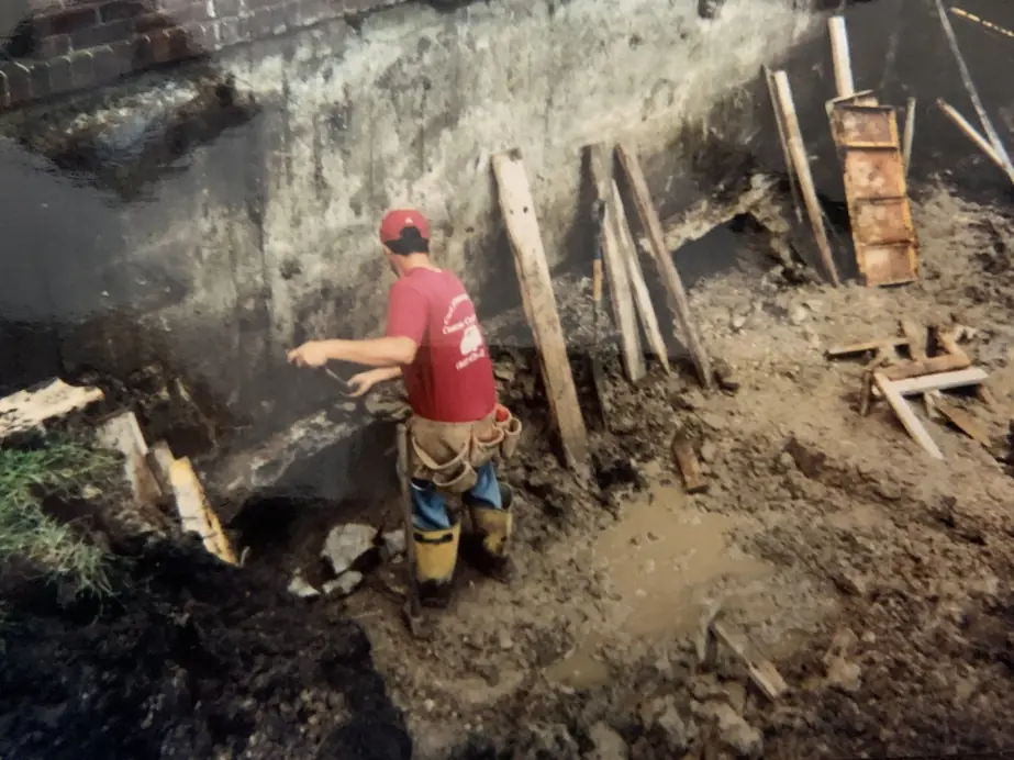 A Carl Johanson & Son crew member in a red company shirt hand-finishing a house foundation wall in an open trench, timber bracing staked along the wall